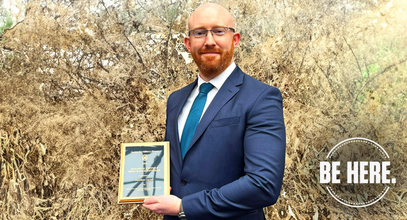 Person in a blue suit holding an award plaque, standing in front of leafless shrubs, with the text "BE HERE" at the bottom right.