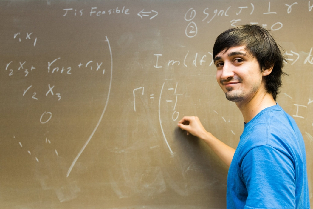Student smiling at the camera as he writes math equations on a chalkboard.