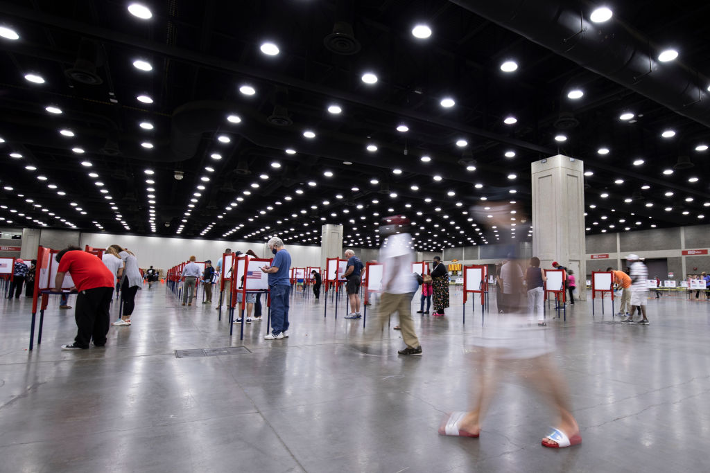 Dozens of people stand at individual polling stations while voting within a large open-concept room