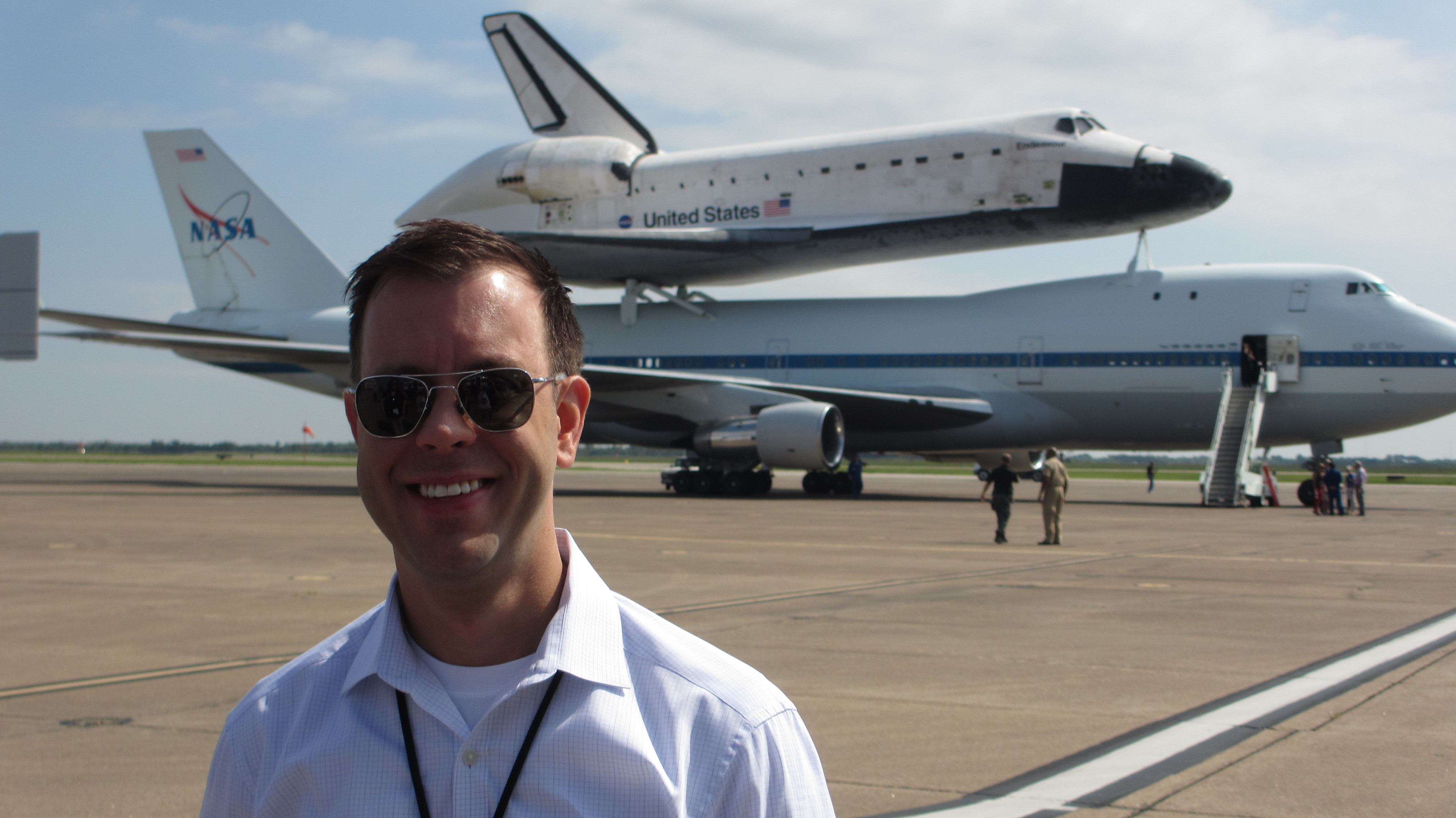 1999 Texas A&M journalism graduate Josh Byerly stands on the tarmac with the Space Shuttle Endeavour atop a NASA Shuttle Carrier Aircraft in the background