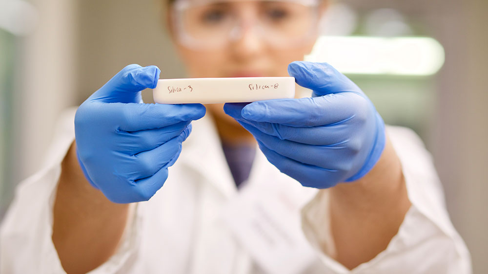 A female researcher wearing goggles, blue gloves and a white lab coat holds a rectangular polymer in her outstretched hands at eye level