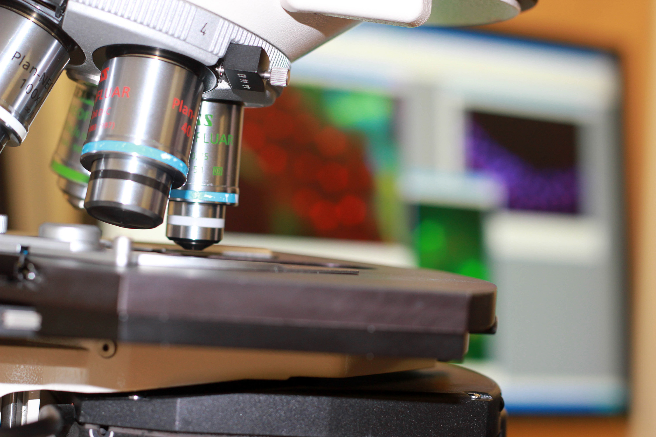 Close up of a microscope's objective lenses, nose piece and stage with a computer monitor featuring science images in the background