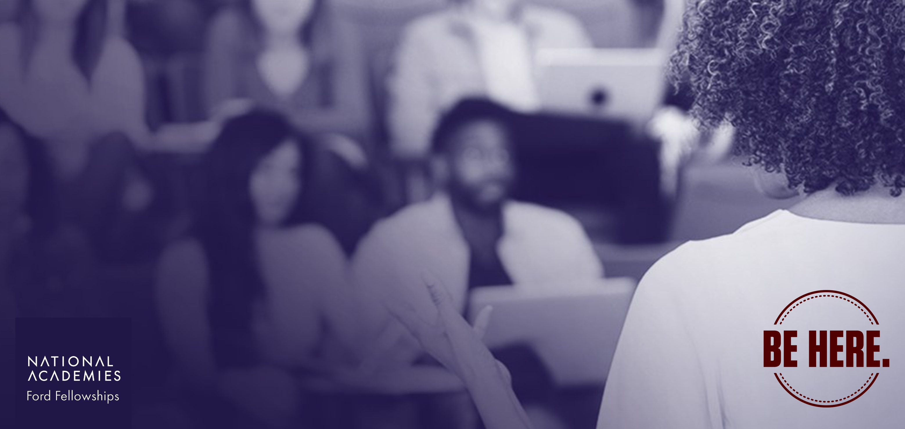 A female presenter with outstretched left hand addresses a multi-race and gendered audience seated in lecture-style classroom