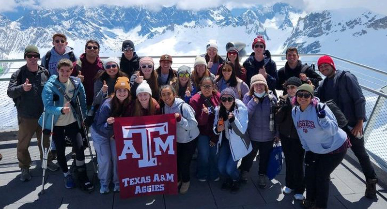 Texas A&M students participating in the 2023 Student Experiences Abroad in Meteorology program pose with a maroon Texas A&M University flag with the Alps in the background