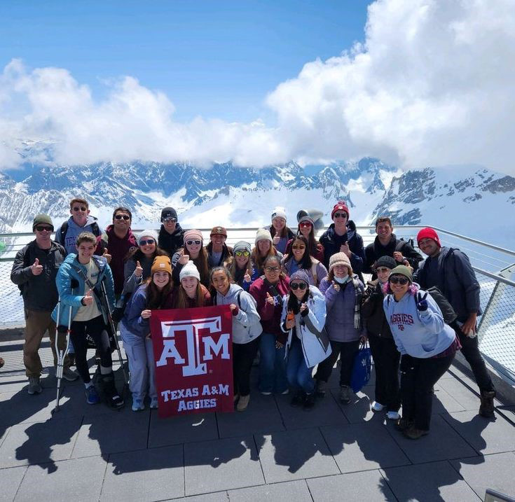 Texas A&amp;M students participating in the 2023 Student Experiences Abroad in Meteorology program pose with a maroon Texas A&amp;M University flag with the Alps in the background