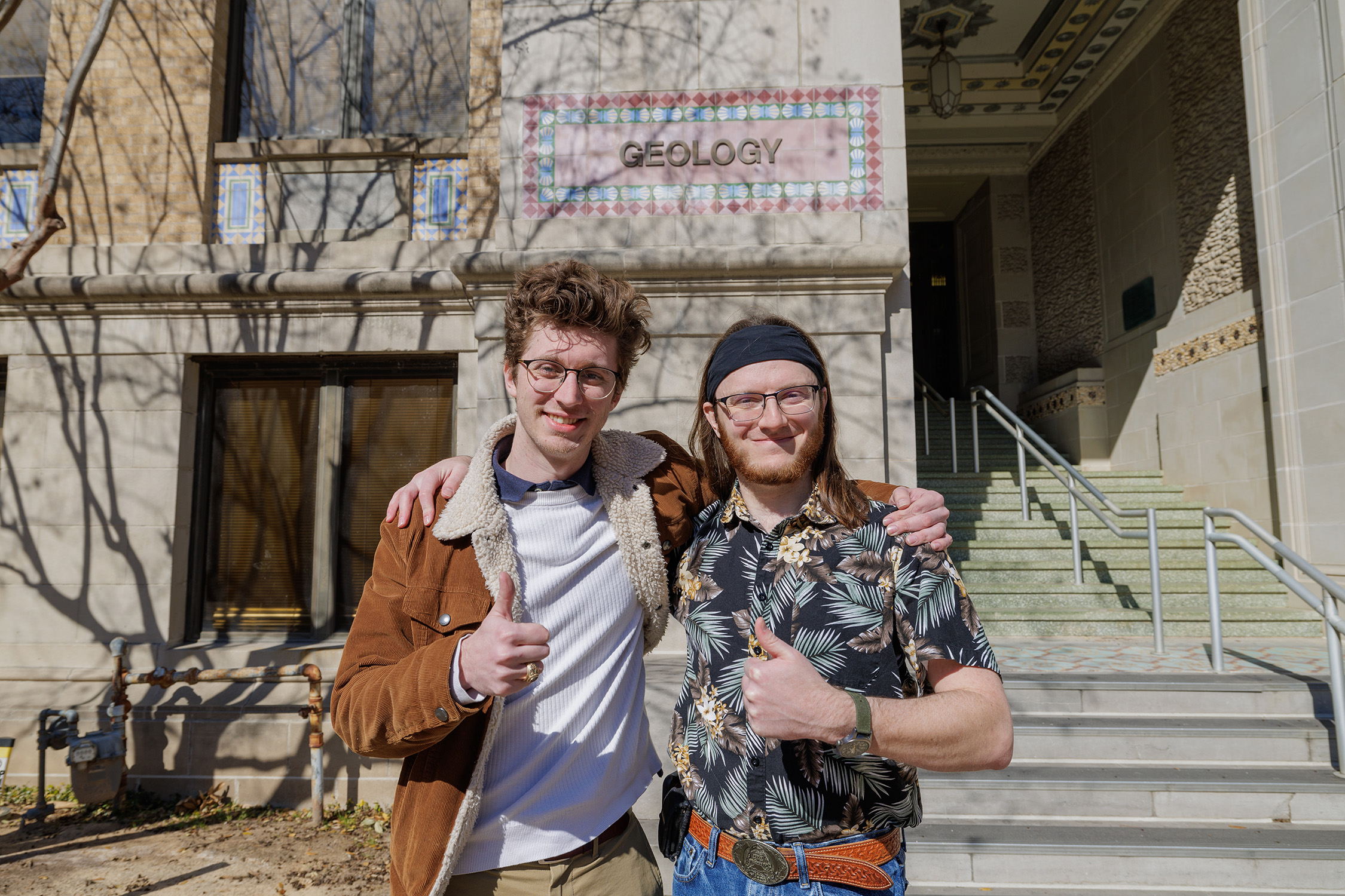 Texas A&M University geology graduate students Dirk van de Laar and Reid "Zeke" Buskirk pose arm in arm while flashing thumbs up signs outside the Halbouty Geosciences Building on the Texas A&M campus