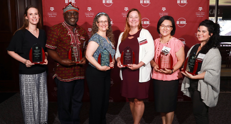 Group photograph of the six recipients of 2024 College of Arts and Sciences Faculty Excellence Awards, posing with their plaques in front of a maroon backdrop featuring the college logo and the "Be Here" stamp in white