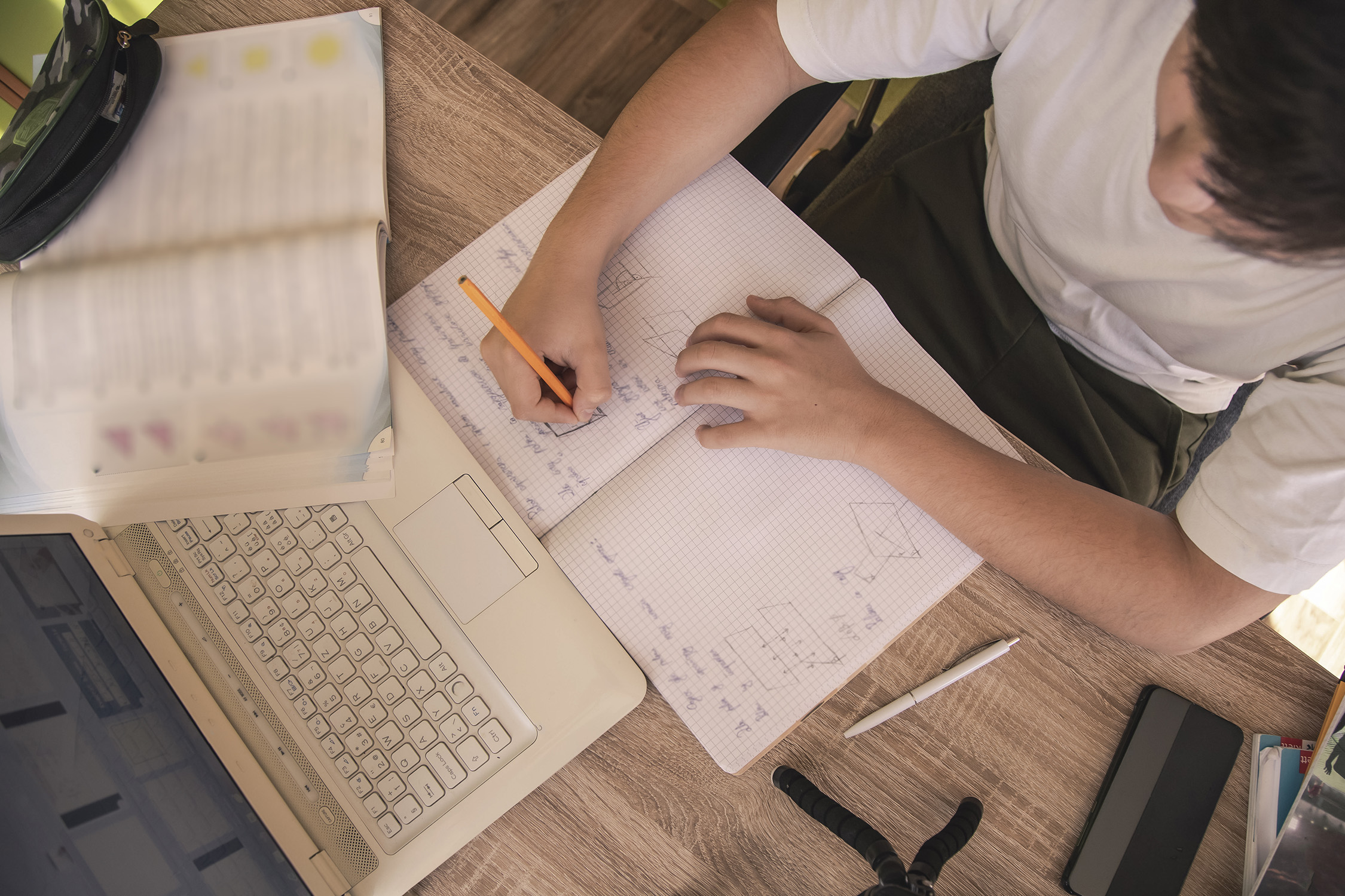 A male student sits at a desk, working on a mathematical problem using a notebook and a laptop