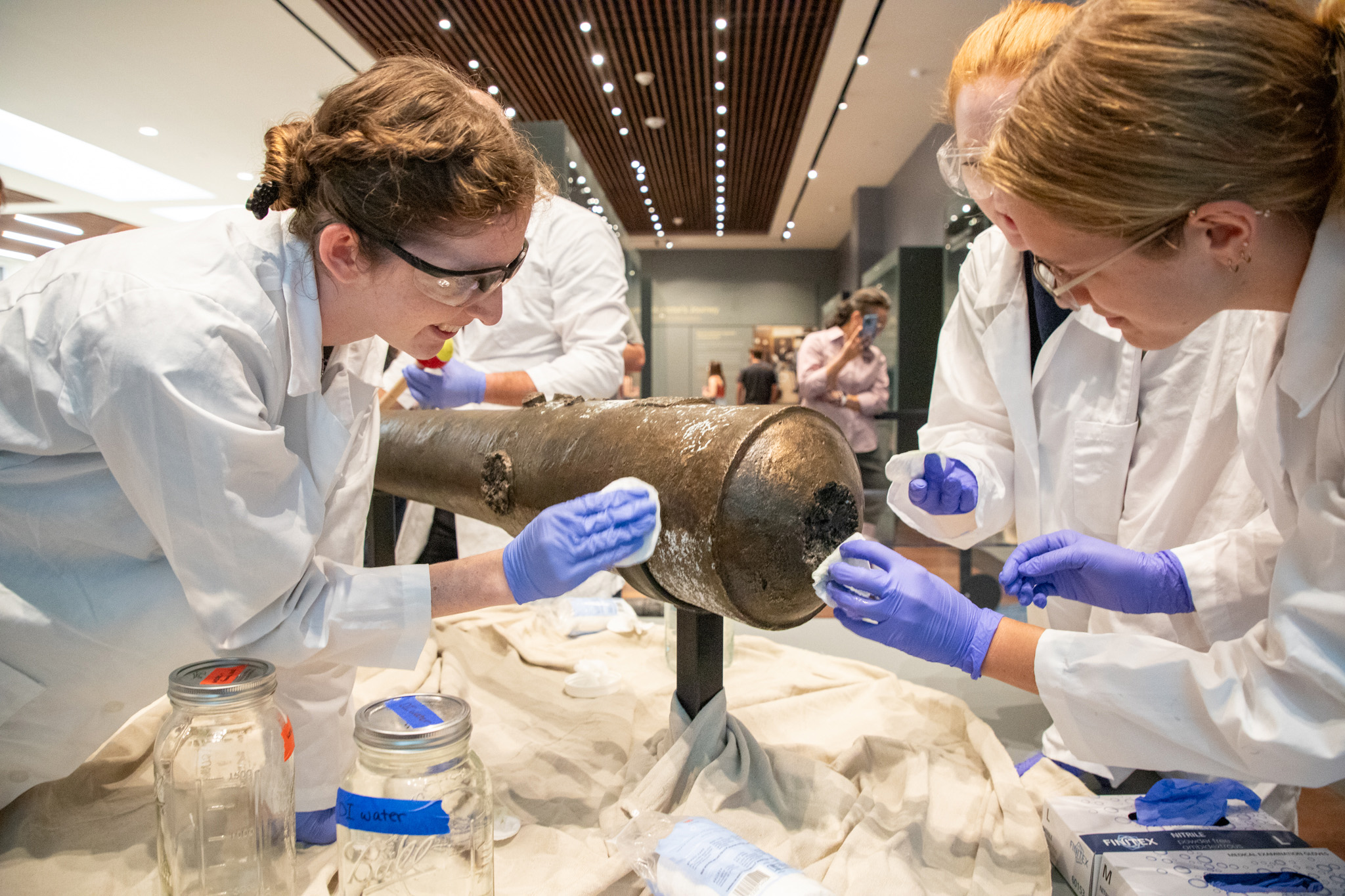 Four female graduate students from Texas A&M University wearing white lab coats, goggles and blue latex gloves work to clean a bronze cannon at the Alamo Museum in San Antonio