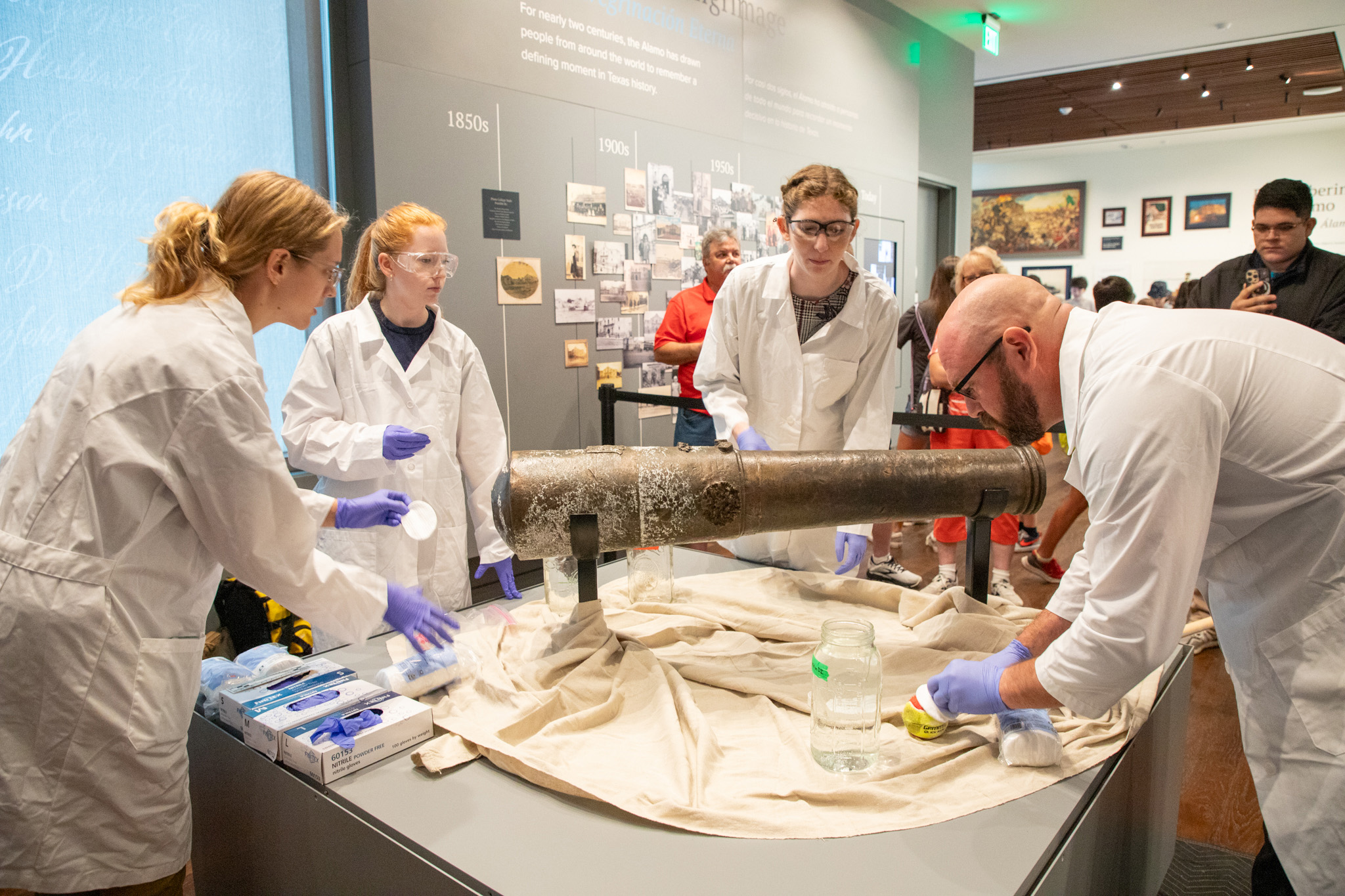 Three female graduate students from Texas A&amp;M University wearing white lab coats, goggles and blue latex gloves work with Texas A&amp;M anthropologist Chris Dostal to clean a bronze cannon at the Alamo Museum in San Antonio