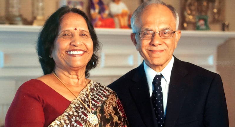 Two individuals smiling for the camera in a home setting, with a photo of Mahatma Gandhi in the background. One person is wearing a sari and the other is dressed in a suit.