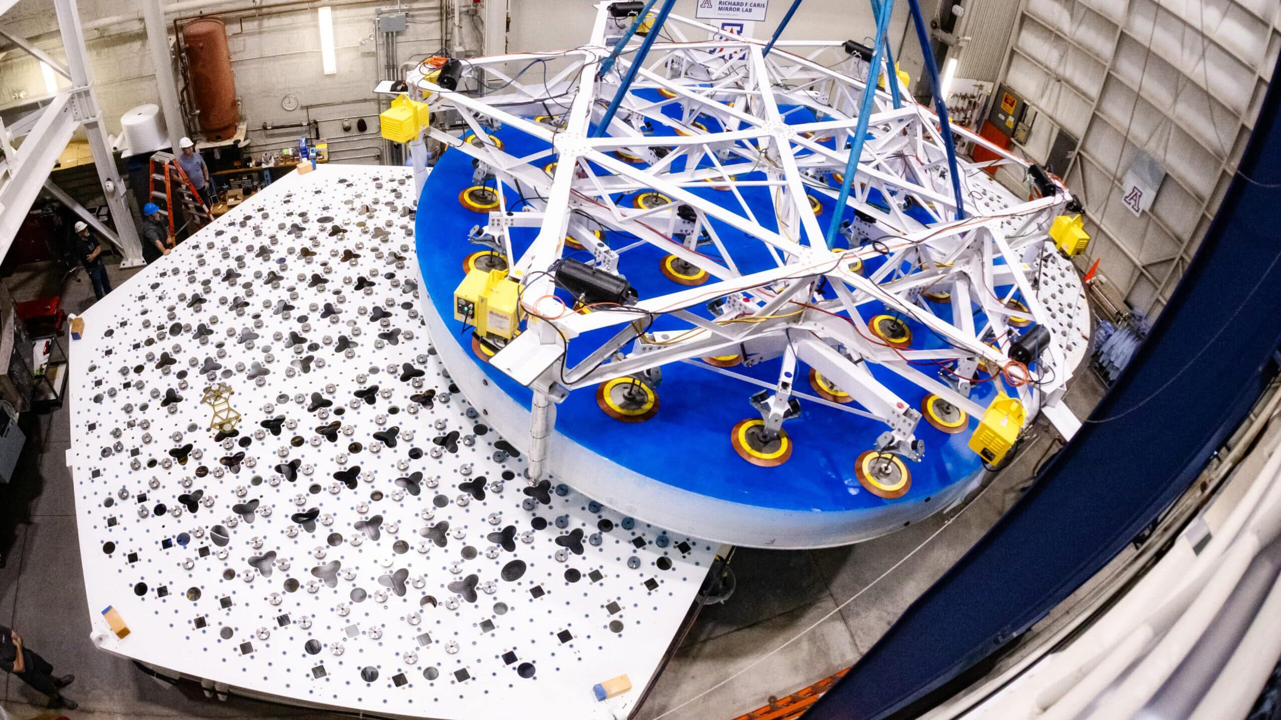 Overhead view of a completed 8.4-meter-diameter primary mirror transport and placement for the Giant Magellan Telescope, suspended above the support system prototype at the University of Arizona’s Richard F. Caris Mirror Lab