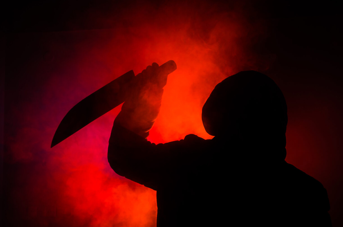 A silhouetted hooded figure holding a machete-type knife poised to strike as smoke swirls against a blood-red background