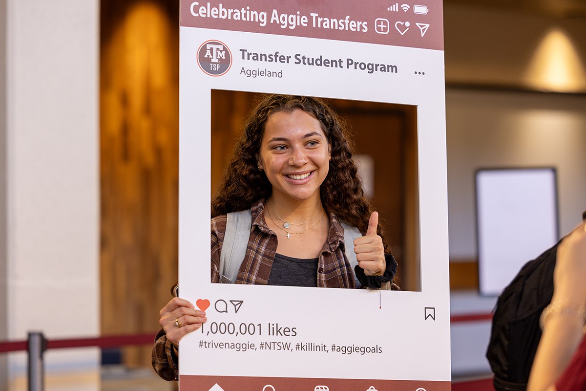 A student smiles for the camera and flashes a gig 'em thumbs up while behind a social media photo booth-style prop during an event for transfer students at Texas A&M University