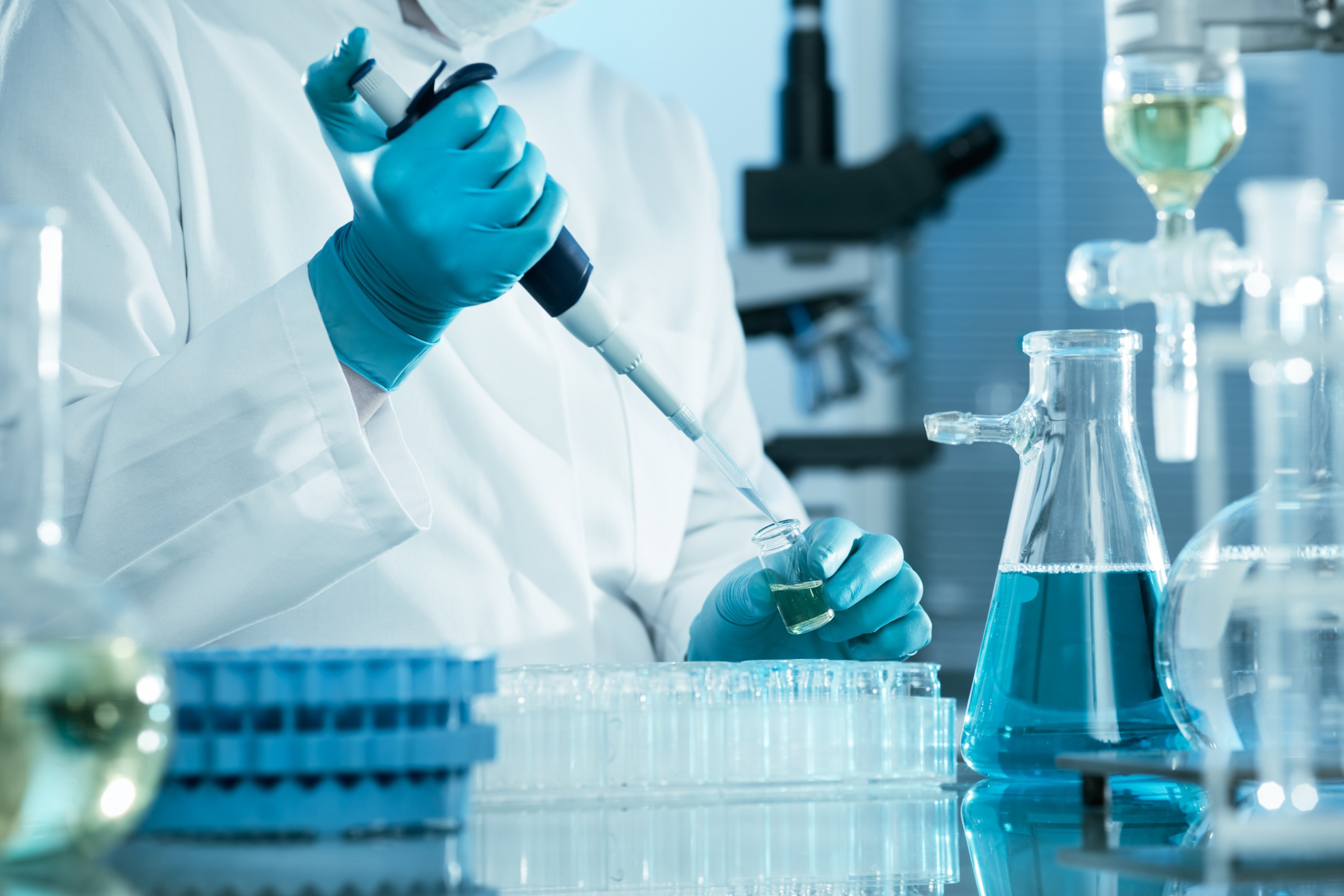 Stock image of a researcher wearing blue latex gloves while pipetting at a laboratory bench filled with glassware, microscopes and other research-related equipment