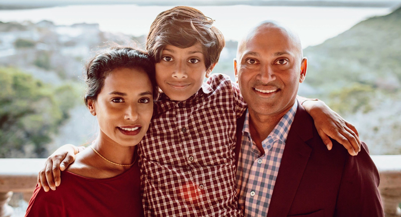 A family of three, featuring two adults and one child, smiling towards the camera with a scenic blurred background of hills and sky.