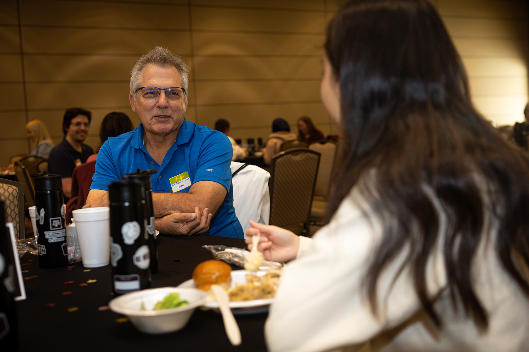 Two individuals conversing over a meal at a dining table, with coffee cups and food plates visible.