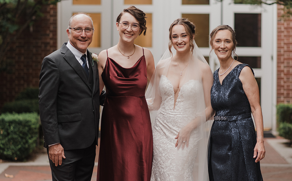 Four individuals in formal wear smiling in a courtyard setting, from left to right: one person in a suit, two in elegant dresses, and another in a dark gown.