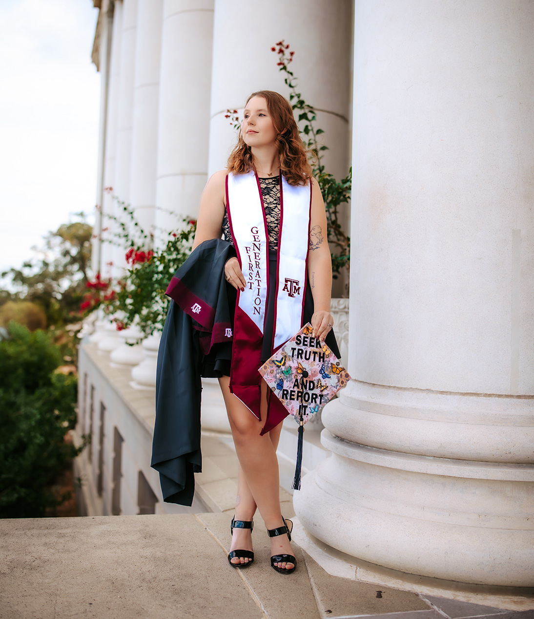 Megan standing confidently in front of the large white columns of the Jack K. Williams Administration Building, wearing a "First Generation" stole, holding a decorated graduation cap that reads "Seek Truth and Report It," and carrying her Texas A&amp;M gown over her arm.