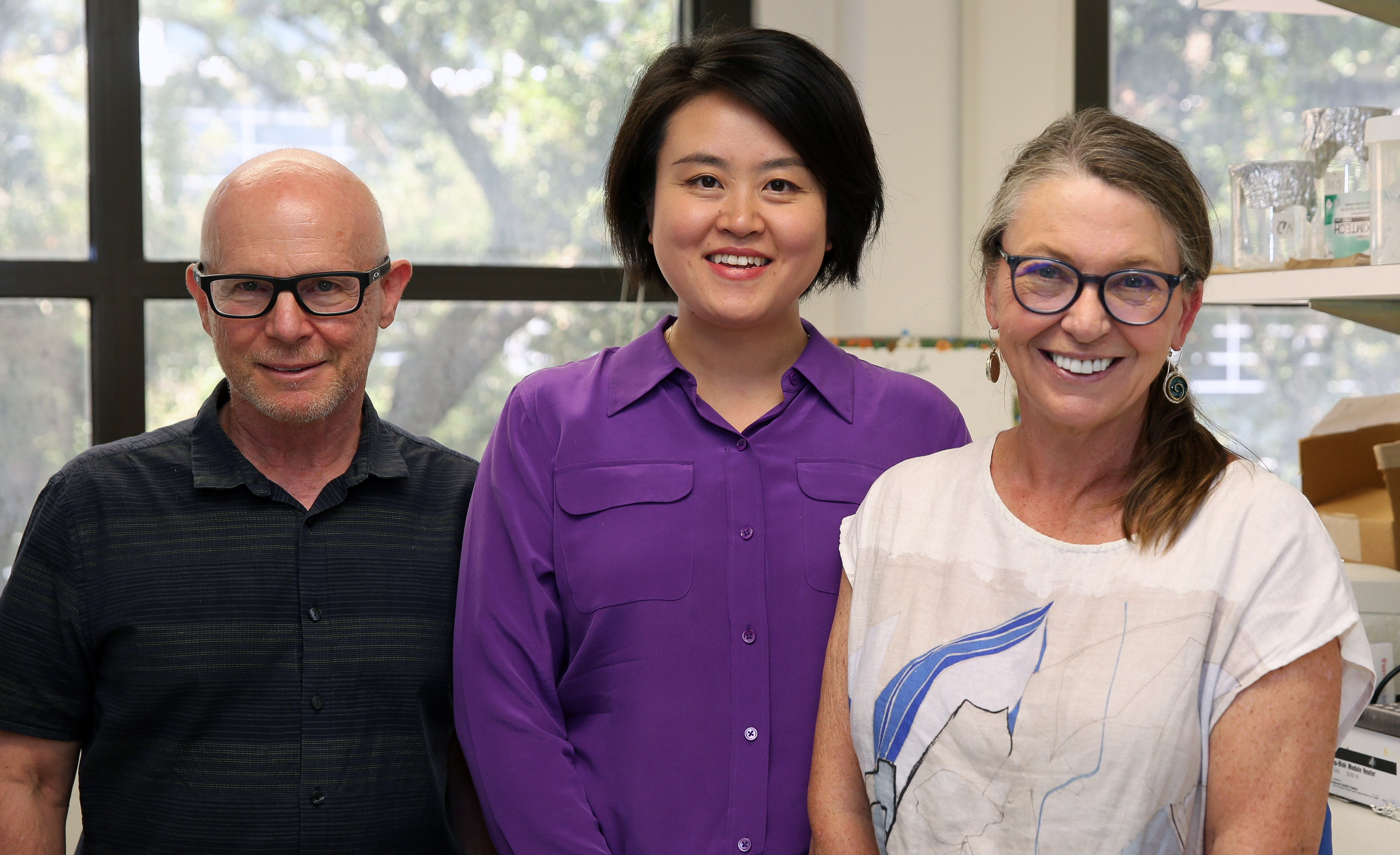 Texas A&amp;M University biologists Dr. Matthew Sachs, Dr. Wanhe Li and Dr. Deborah Bell-Pedersen are smiling in an office setting, with Dr. Li in a purple shirt flanked by two others, Dr. Sachs wearing glasses and a black shirt, and Dr. Bell-Pedersen in a white shirt with a blue abstract design.