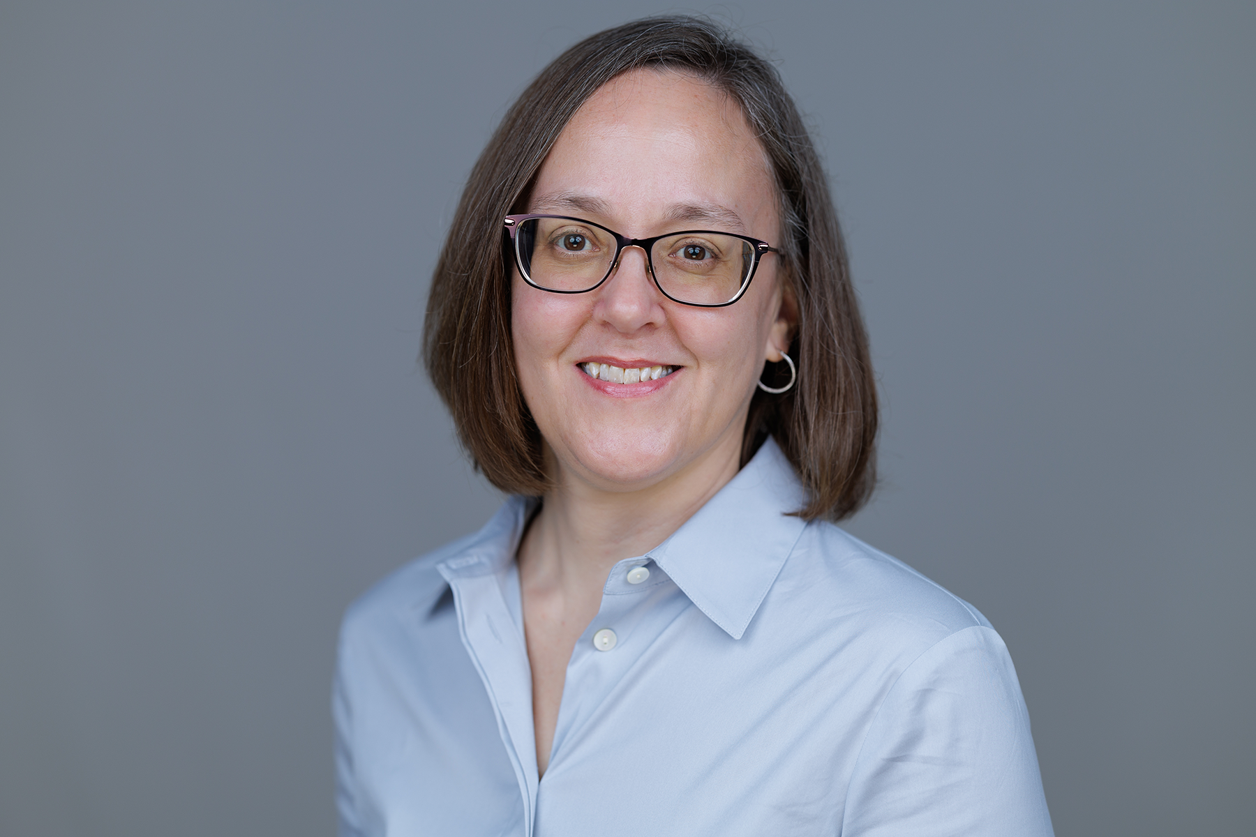 Professional headshot of Dr. Kristi Sweet with glasses, wearing a light blue shirt, set against a gray background.