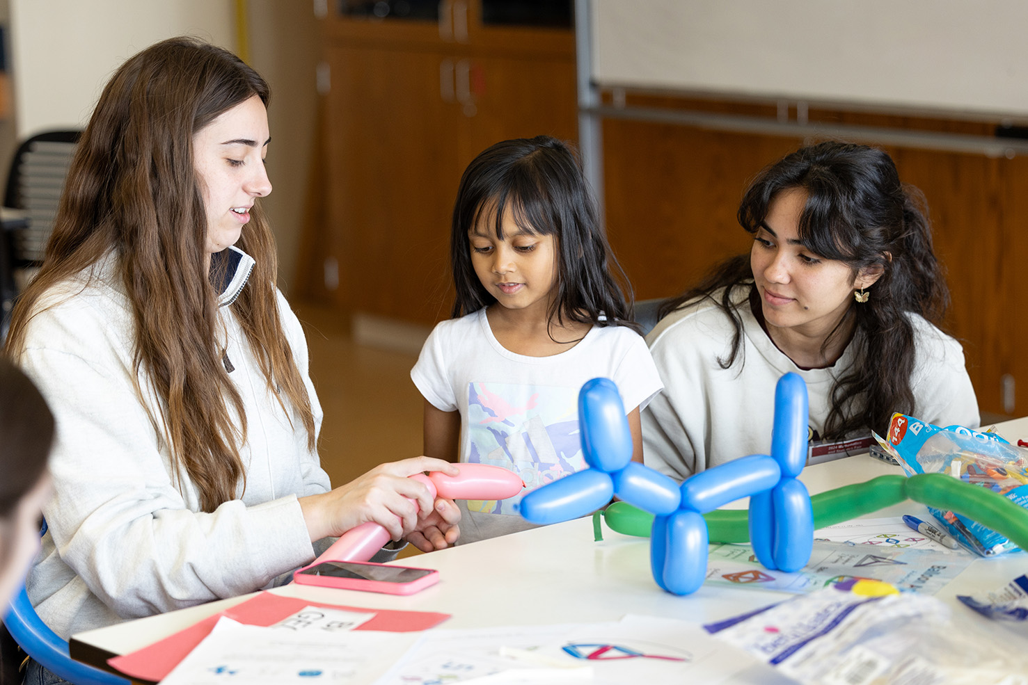 An undergraduate student volunteer shows a young participant and another undergraduate volunteer how to twist balloons to make balloon animals. Another balloon animal shaped like a dog is perched up on a table beside them.