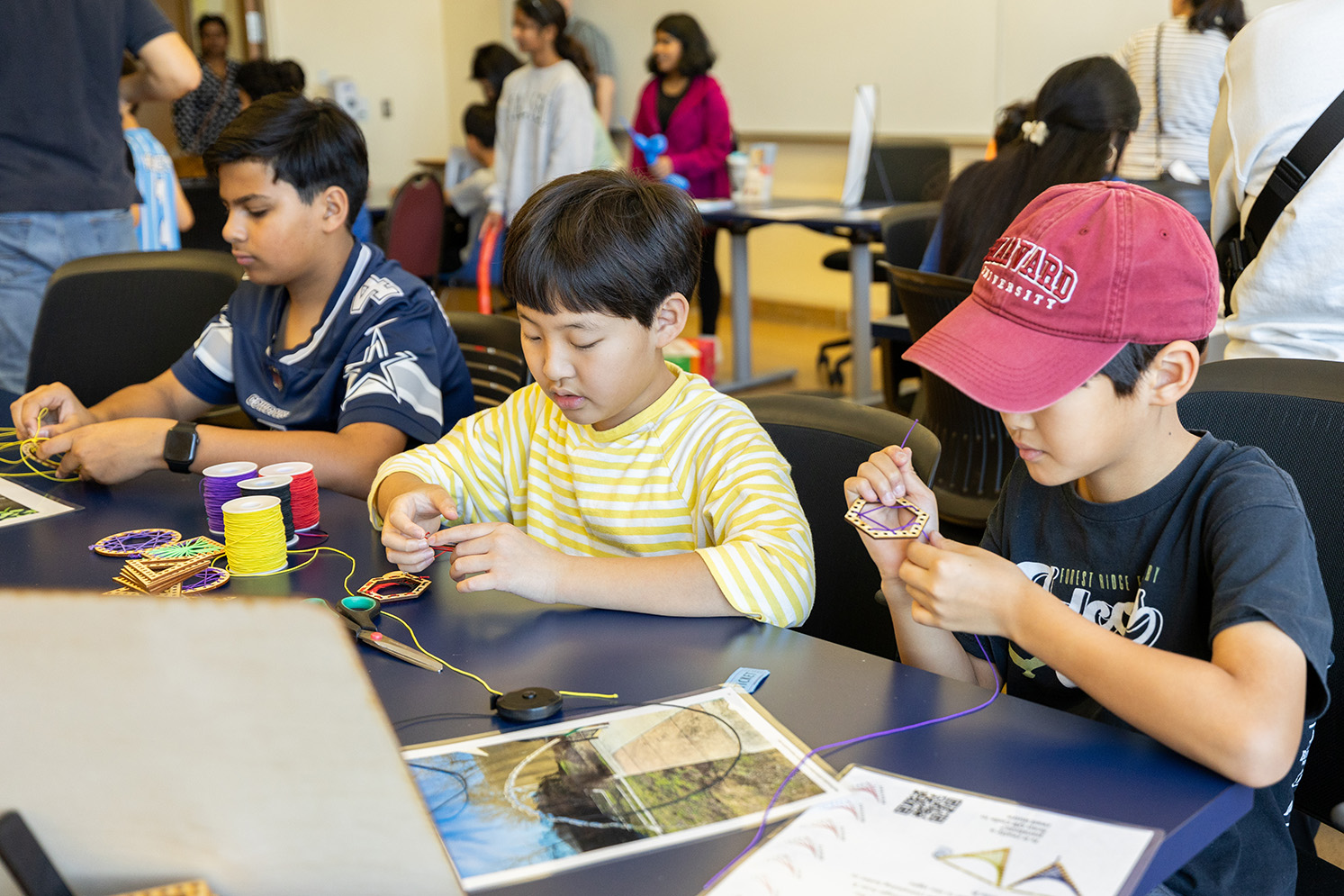 Three young Fair participants sit at a table following mathematical instructions to weave colorful thread through pre-cut holes through a small wooden border of a hexagon. Spools of colorful thread are set on the table in front of them, along with completed weaves and other mathematical illustrations. The background is filled with other Fair participants engaging with other Fair activities.