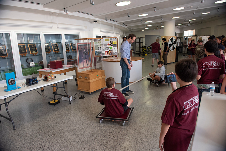 A person stands addressing a group of students wearing maroon shirts labeled "CENTAUR" in a bright classroom with educational exhibits and posters around. Some students sit on the floor while others are on rolling boards.