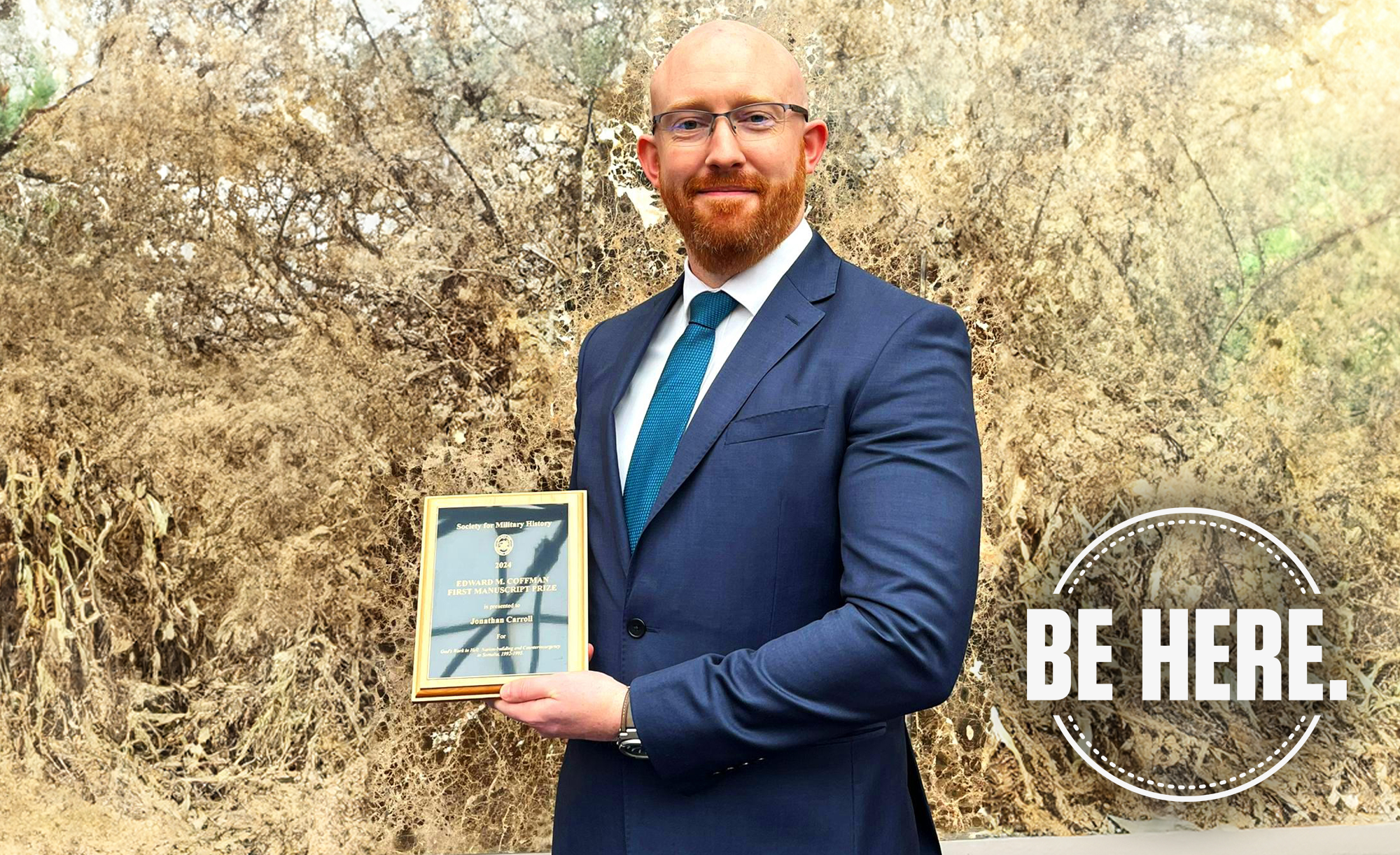 Person in a blue suit holding an award plaque, standing in front of leafless shrubs, with the text "BE HERE" at the bottom right.