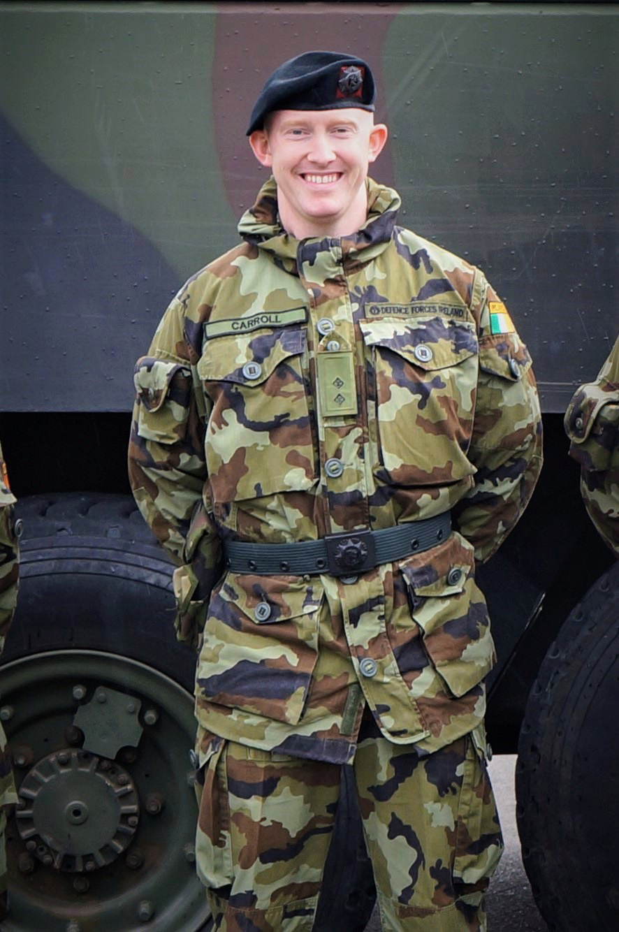 Carroll stands in front of a military tank wearing military fatigues, smiling.