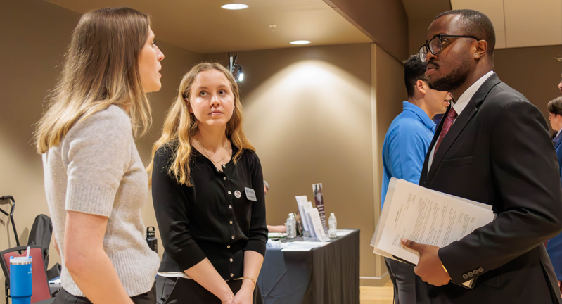 Two representatives from Trenegy speak with an Economics student dressed in a suit and carrying a folder.