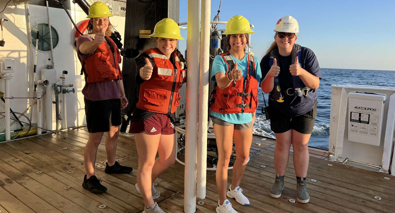 Four individuals wearing safety gear including helmets and life jackets, giving thumbs up on the deck of a ship at sea.