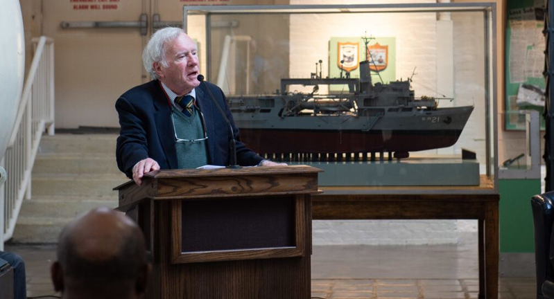 Don Walsh speaking at a podium in a museum with a large ship model in a display case in the background.