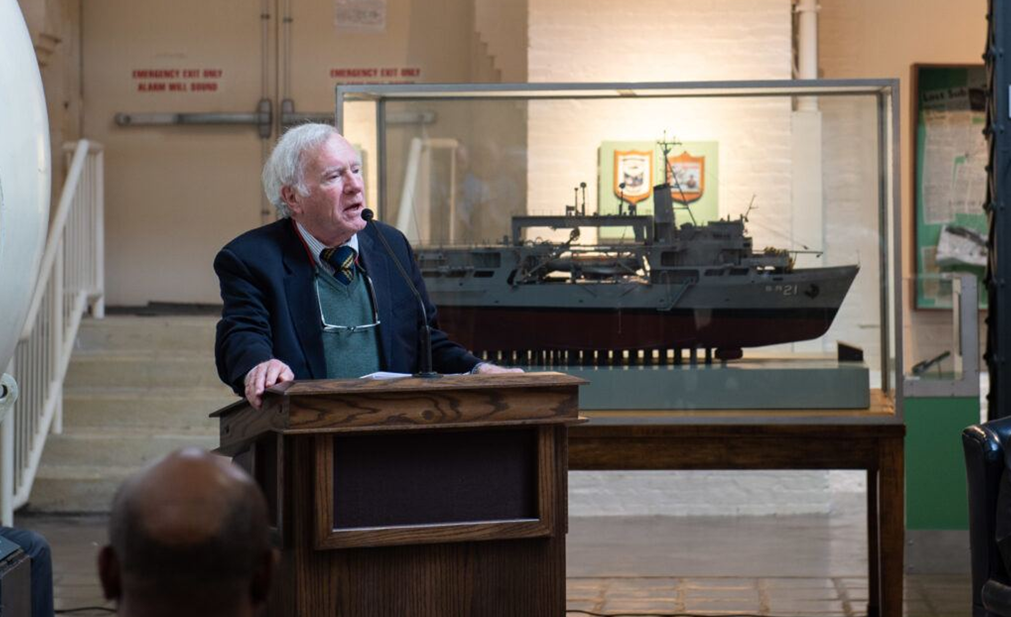 Don Walsh speaking at a podium in a museum with a large ship model in a display case in the background.