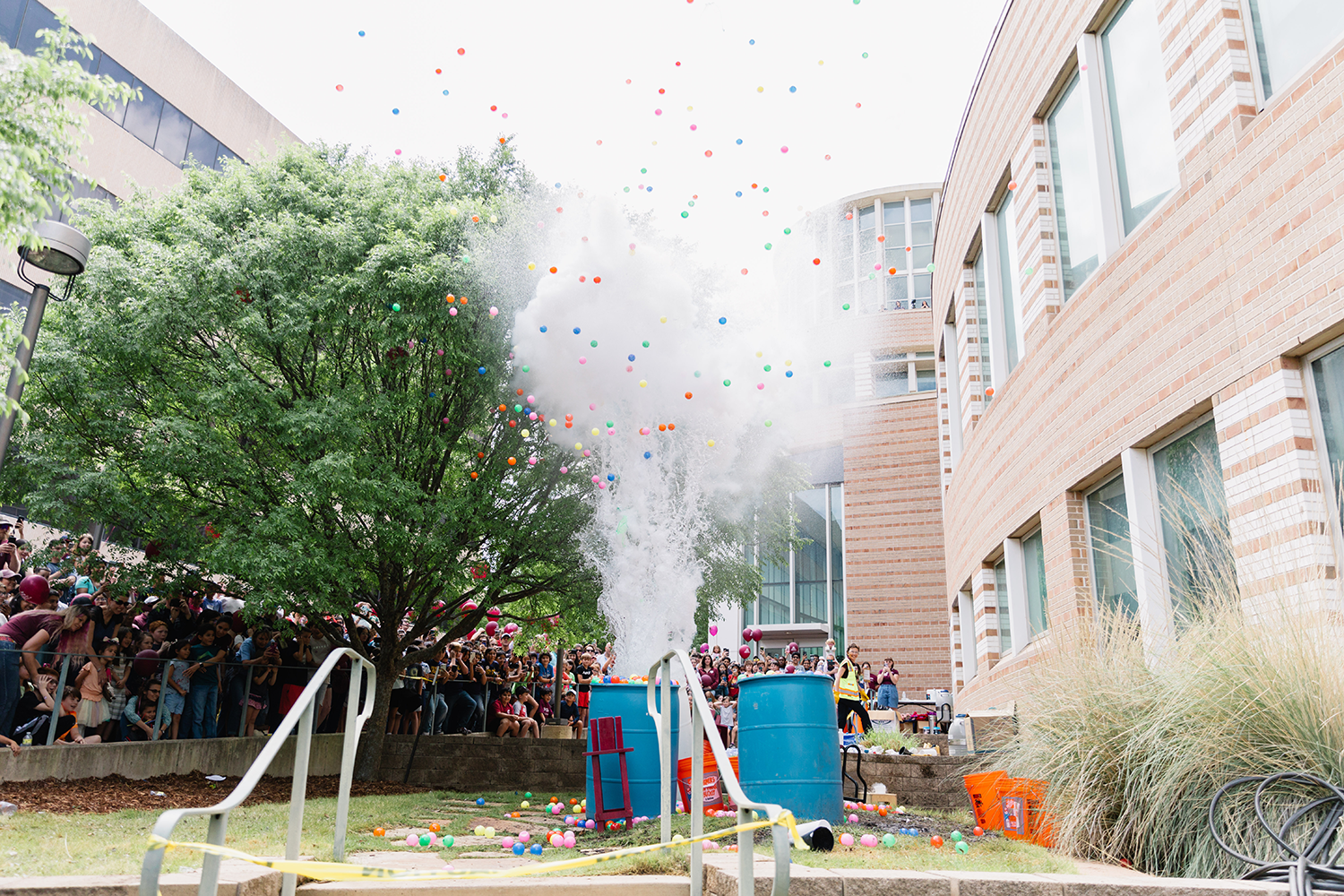 A lively 2023 Physics Festival at Texas A&amp;M University, where a crowd is gathered with balloons emerging explosively from blue barrels amidst a campus setting.