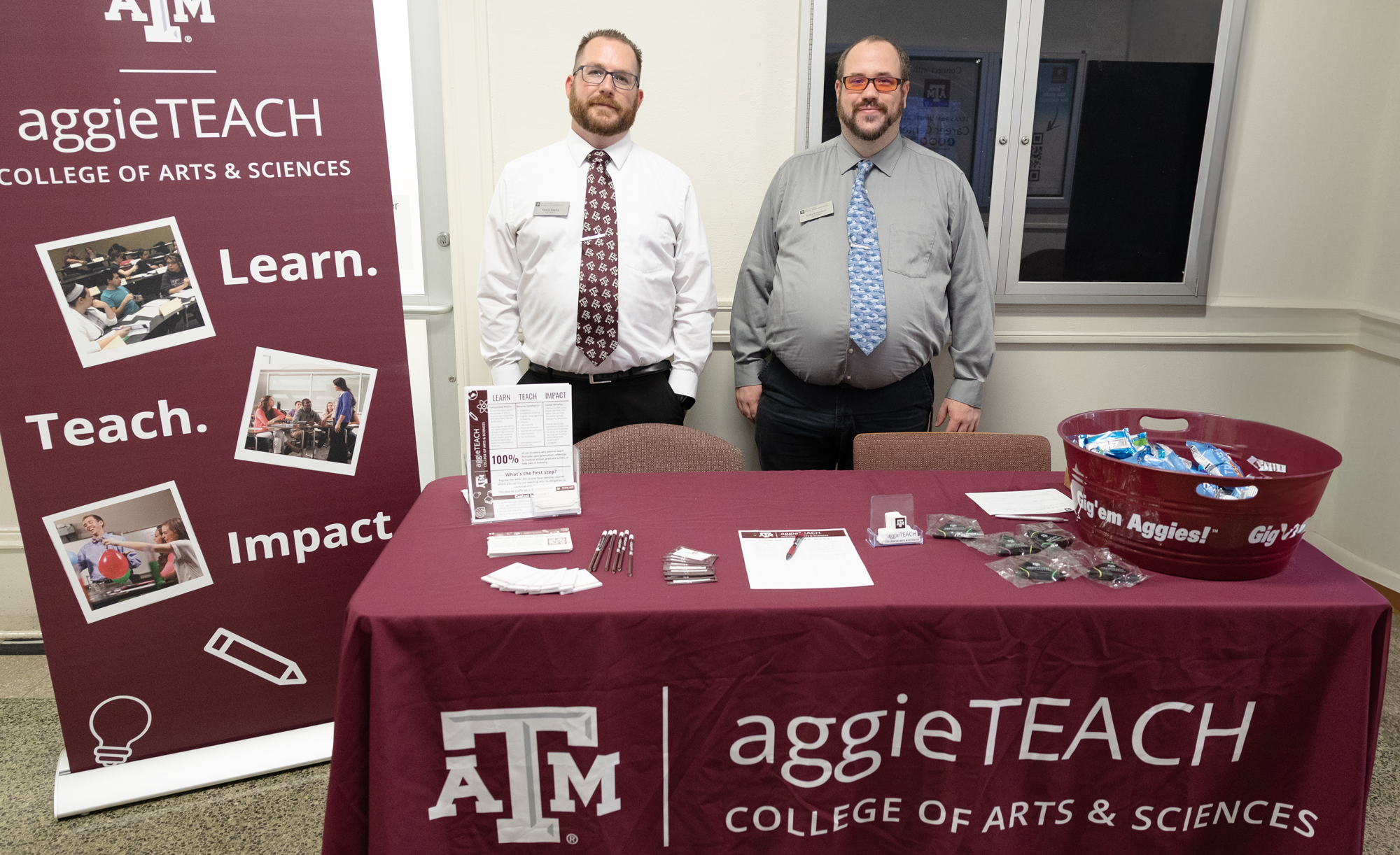 Kenric Davies and Colby Richardson standing behind a promotional table for the AggieTEACH program at Texas A&amp;M University, flanked by banners displaying the university's logo and the College of Arts &amp; Sciences. The table features brochures and promotional items.