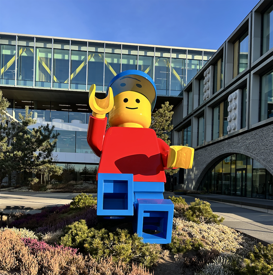 A large LEGO figure stands in front of a modern building with glass facades and surrounding greenery.