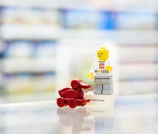 A LEGO minifigure standing next to a small LEGO car, with "LEGO House" printed on the minifigure's torso. The background features a blurred view of a store shelf filled with products.