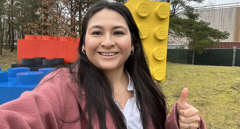 Jennifer Chavez smiling with a thumbs up in front of large LEGO bricks.