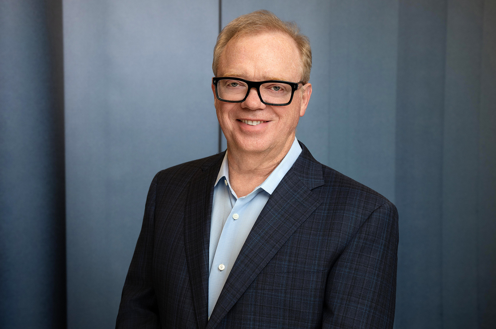 Portrait of 1987 Texas A&M University graduate Anthony Wood, who stands against a blue-toned backdrop wearing thick-rimmed black glasses, a light blue button-down dress shirt and a navy blue plaid sport coat.