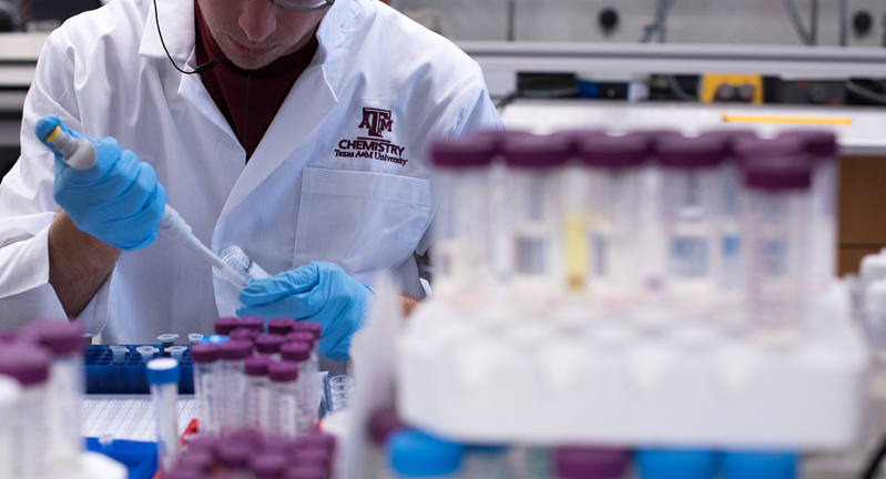 A Texas A&M University lab technician in a white lab coat labeled "Chemistry" is pipetting a substance into a test tube, surrounded by racks of samples.