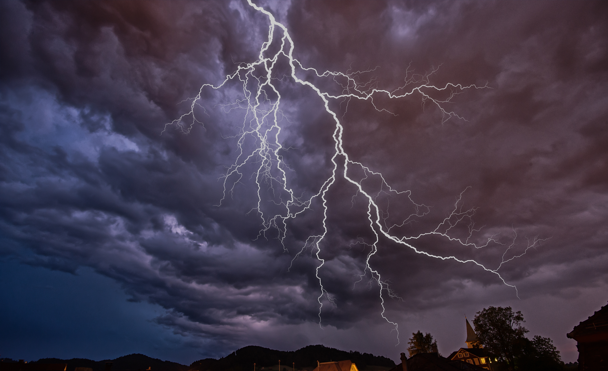 Lightning striking over a landscape with mountains and houses under a dramatic cloudy sky.