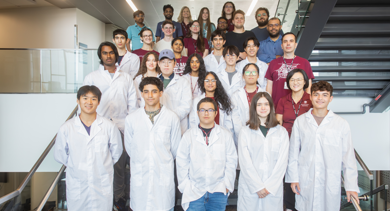 Group of high school students in white coats standing in a staircase along with faculty and staff from Texas A&amp;M's Chemistry Department.