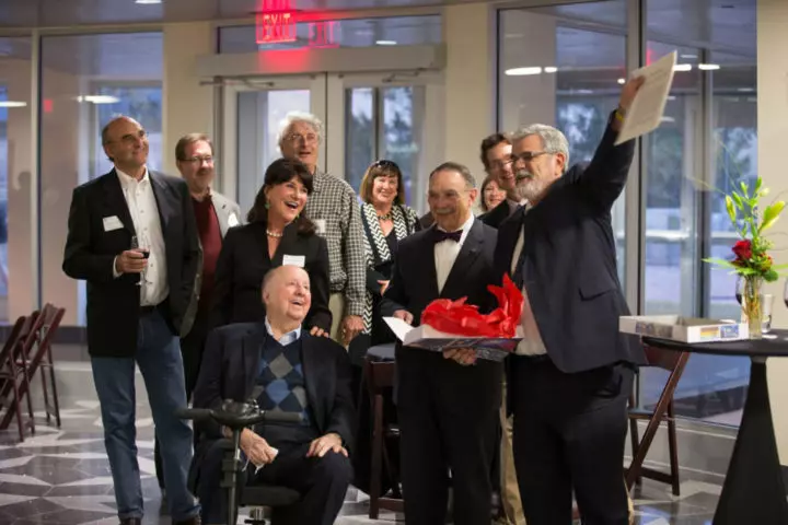 A reception with a donor with Physics and Astronomy faculty and university leadership. Everyone is smiling and appears excited. A man in a suit is holding up a paper that everyone is looking at.